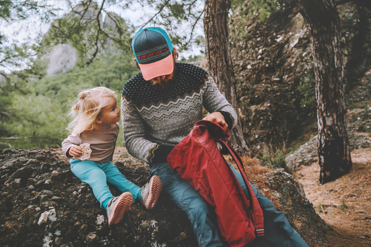 Ett barn sitter på en sten och äter en riskaka med hennes pappa plockar ut något ur ryggsäcken.