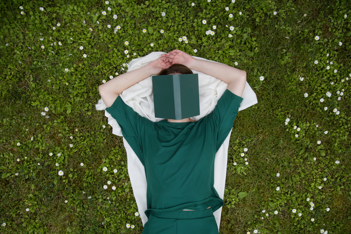 En person ligger på gräset. Gräset blandas med vita små blommor. Personen har somnat med en bok i ansiktet.
