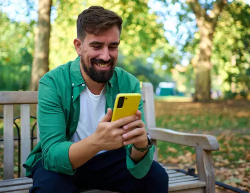 En man i grön skjorta sitter på en parkbänk och kollar sin mobiltelefon.
