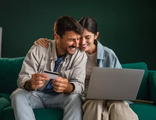 Ett par sitter i soffan och kollar på en laptop och håller ett kreditkort