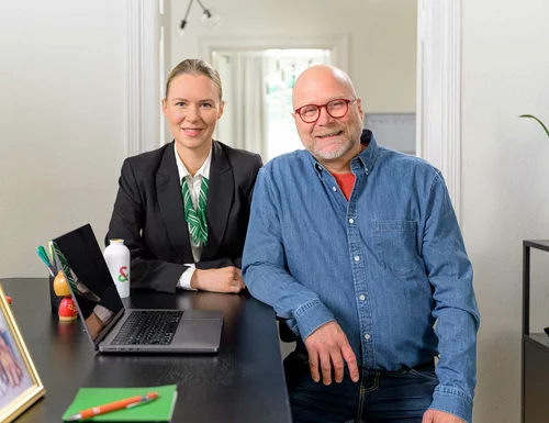 En man och en kvinna står på ett kontor och tittar in i kameran och ler.
