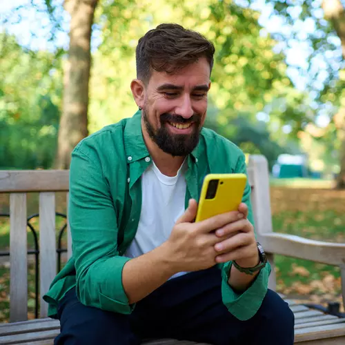 En man med grön skjorta sitter på en bänk och kollar sin mobiltelefon.