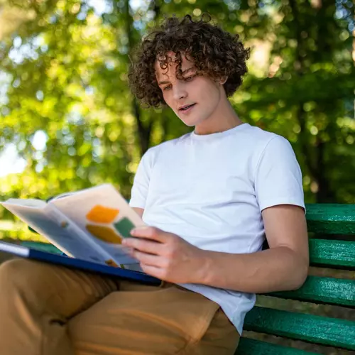 En man sitter på en grön bänk och läser en bok.