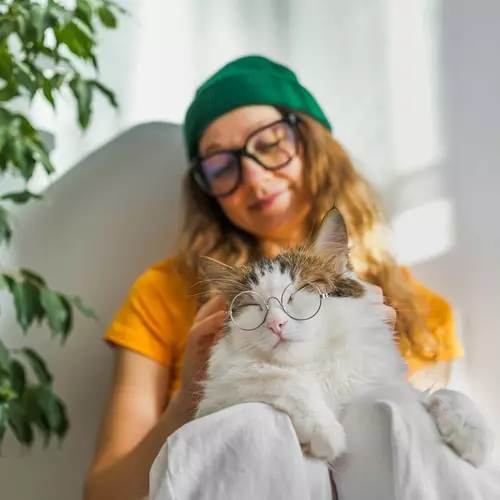 En tjej i grön mössa sitter med sin katt. Katten har glasögon på sig.