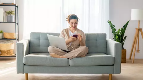 En kvinna sitter hemma i soffan med sin mobil och laptop